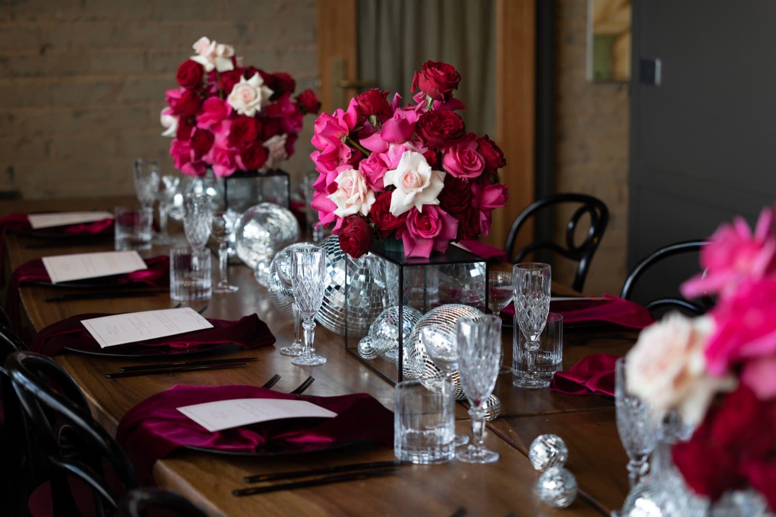 A long table dressed for a wedding: pink and red roses, crystal glassware, burgundy velvet napkins and mirror balls down the middle